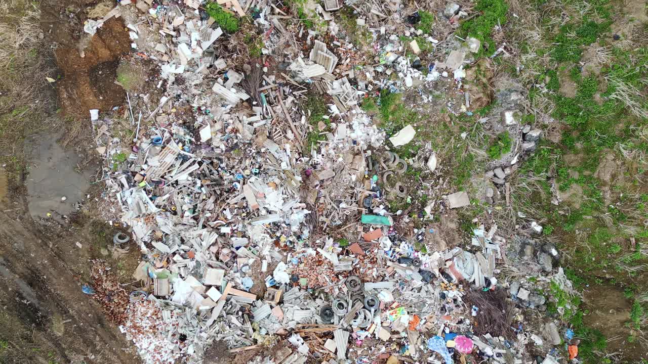 Аэросъемка показывает разбросанный мусор на травянистой местности. Кучи отходов содержат пластик, бумагу и сломанные предметы. Грязная земля окаймляет хаотичную груду мусора. Людей на снимке нет.