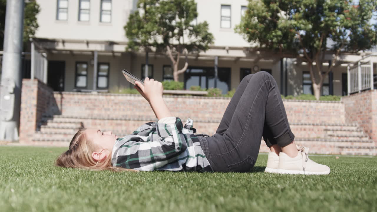Young girl relaxing on grass outside school, reading tablet under sunny sky