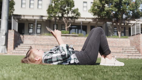 young girl relaxing on grass outside school, reading tablet under sunny sky