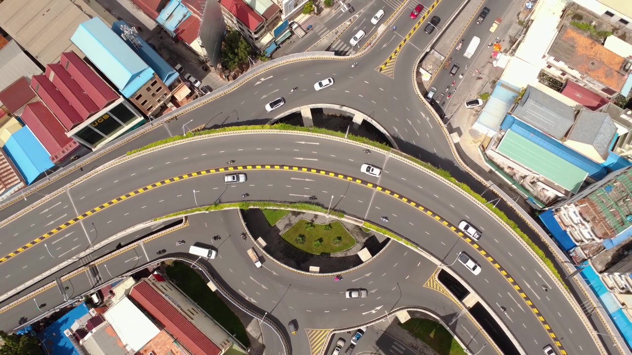 Top-down drone view of a multilevel flyover interchange in Phnom Penh, Cambodia, with moving traffic, sweeping ramps and dense urban development surrounding the roadway.