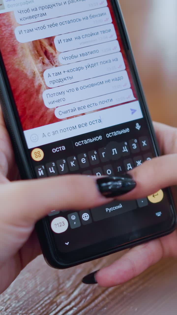 primer plano de un teléfono móvil negro utilizado por una mujer para conversar en idioma ruso en la pantalla con los dedos escribiendo en el teclado, mesa de madera con efecto de luz borrosa vibrante alrededor