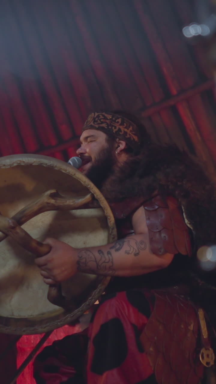 Tattooed man with shamanic drum sings traditional ritual songs during performance in old local house. Musician uses folk instrument at show