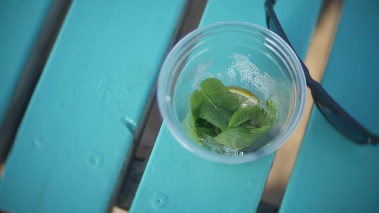 Cup holds mint leaves, lemon slices and melting ice on a bright turquoise picnic table. Overhead close-up shows a refreshing drink remnant beside black sunglasses