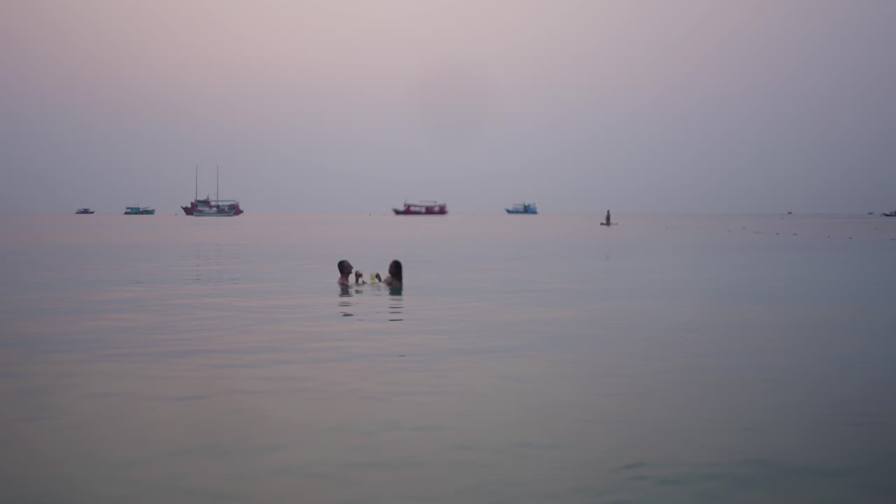People swimming in the ocean with boats and a paddleboarder at sunset