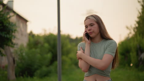 young girl appears exhausted and frustrated during phone call, walk pass a pole by the side while talking, set against a blurred outdoor background of greenery and distant buildings