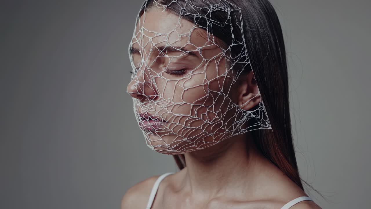 Serene fashion model wearing intricate white mesh mask, standing with closed eyes against neutral gray background, embodying contemporary elegance and artistic design