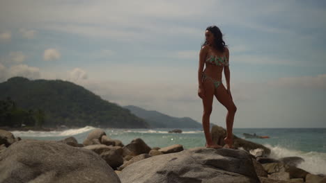 Beautiful-young-woman-in-a-bikini-standing-on-a-rocky-shore-of-an-island-paradise-with-the-waves-crashing-in-slow-motion