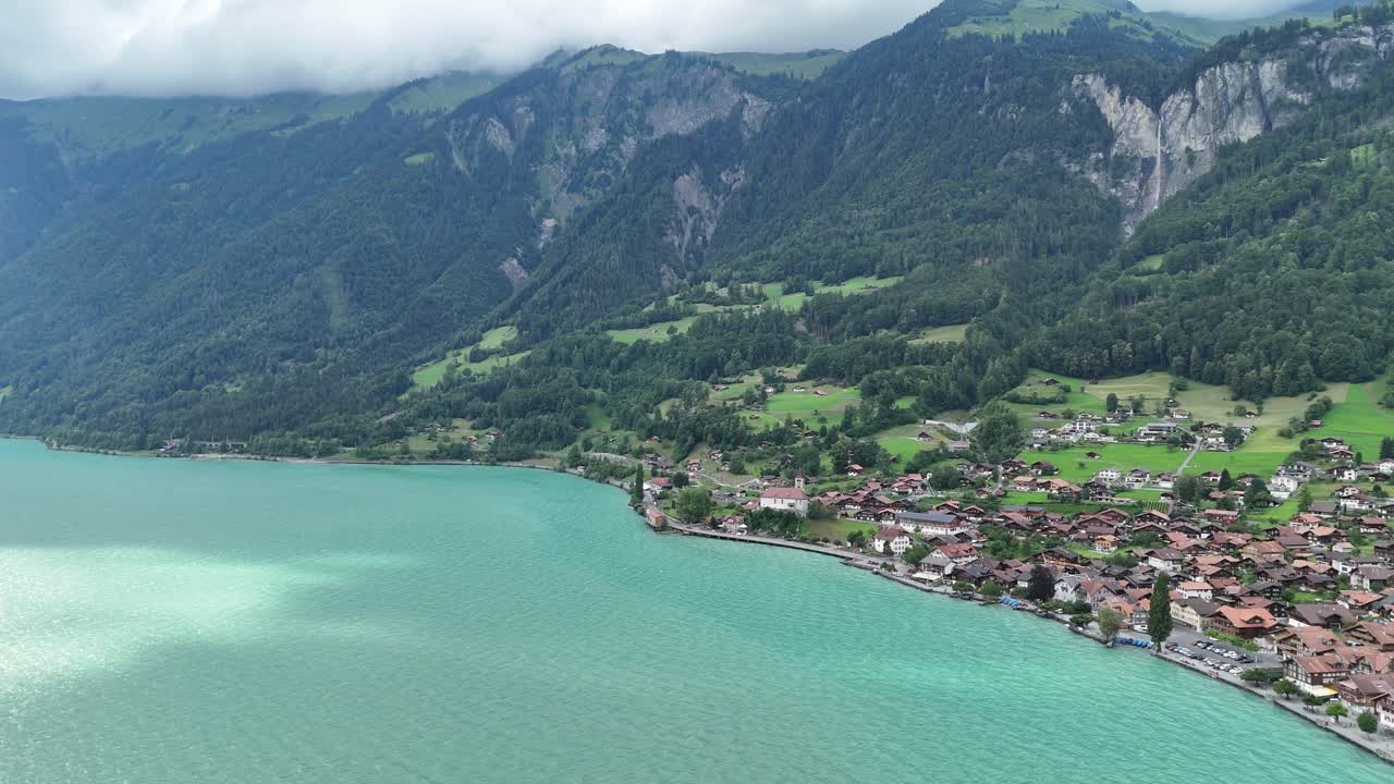 ドローンがターコイズブルーのブリエンツ湖上空を降下し、曇り空の下、魅力的なスイスの村、緑豊かなアルプスの山々、そして流れ落ちる滝が現れる