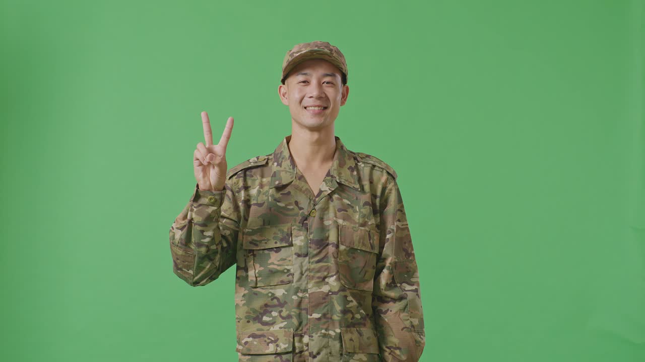 Asian Man Soldier Smiling And Showing Peace Gesture To Camera While Standing In The Green Screen Background Studio