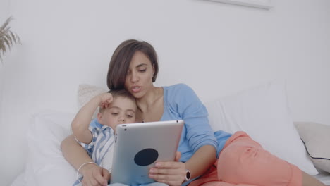 En-El-Dormitorio-Blanco,-Mamá-E-Hijo-Miran-La-Pantalla-De-La-Tableta-Y-Se-Ríen.-Familia-Feliz-En-La-Cama-Por-La-Mañana-Leyendo-Un-Libro-Y-Haciendo-Clic-En-El-Panel-Táctil-De-La-Computadora.