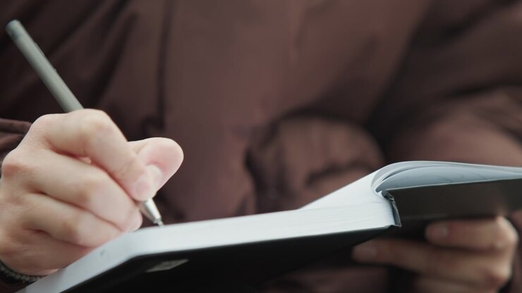 Closeup hands writing in notebook on bench, brown coat, silver pen, overcast light, reflective mood freelance writer drafting notes, steady strokes, page texture visible, shallow depth of field
