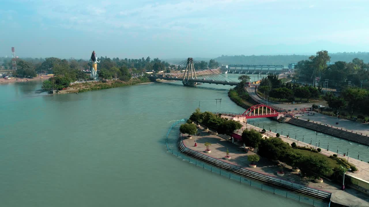 amplia toma aérea de la ciudad sagrada haridwar que muestra la gran estatua del dios hindú lord shiva, el agua que fluye azul brillante del río ganges, el puente sobre el río sagrado ganges y el tráfico que pasa por el puente