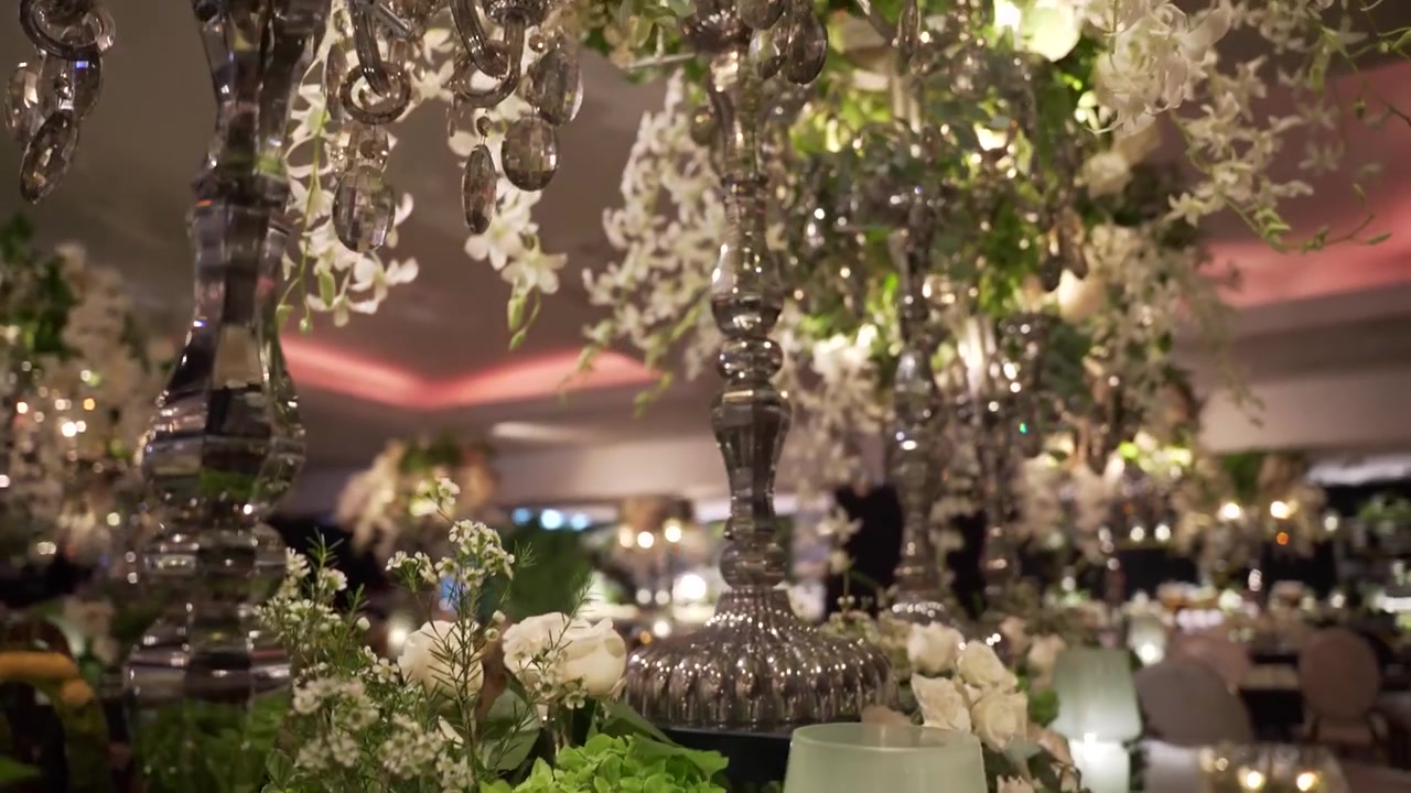 Primo piano stabilizzato di eleganti composizioni floreali e lampadari di cristallo che decorano una location di lusso per ricevimenti di matrimonio con calda illuminazione ambientale.