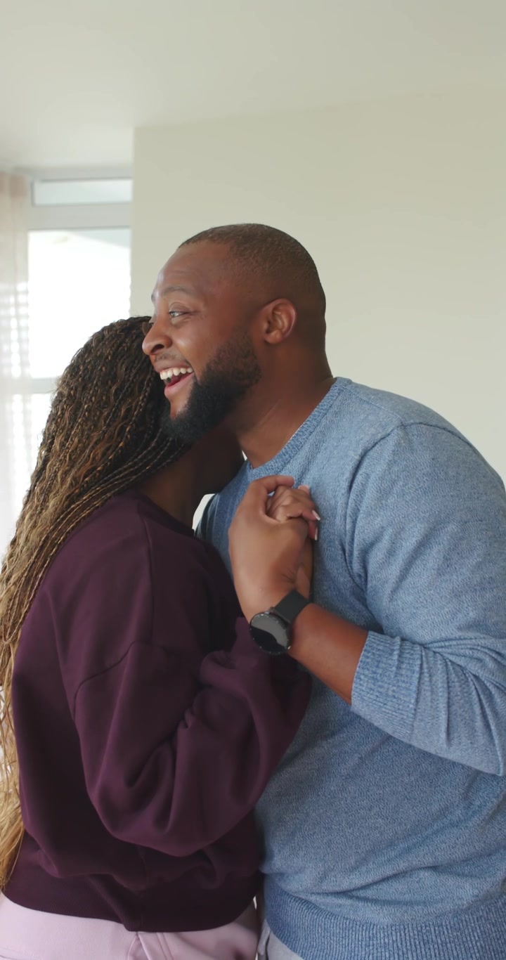 Vertical video: African couple laughing, leaning in and embracing by window, showing ring and watch
