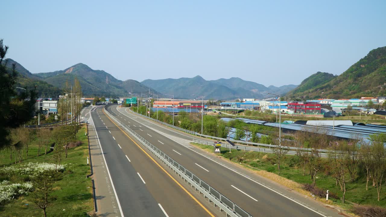 Modern asphalt highway stretches through a scenic mountain valley with one vehicle visible near colorful industrial buildings and agricultural plastic greenhouses in South Korea