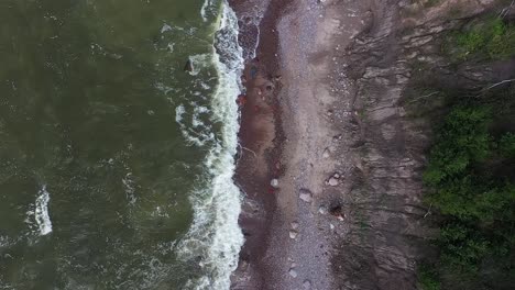 A-top-down-view-from-a-drone-showcases-the-dramatic-high-cliff-coast-on-the-Baltic-Sea-in-Lithuania,-portraying-sizable-waves-and-the-atmospheric,-green-colored-waters