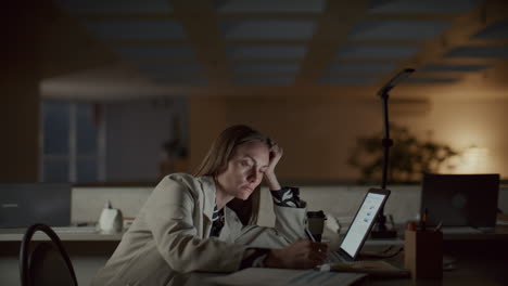 tired businesswoman using laptop and taking notes while overworking at night