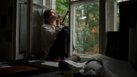Serious-woman-smoking-indoors.-Contemporary-painter-relaxing-on-windowsill.