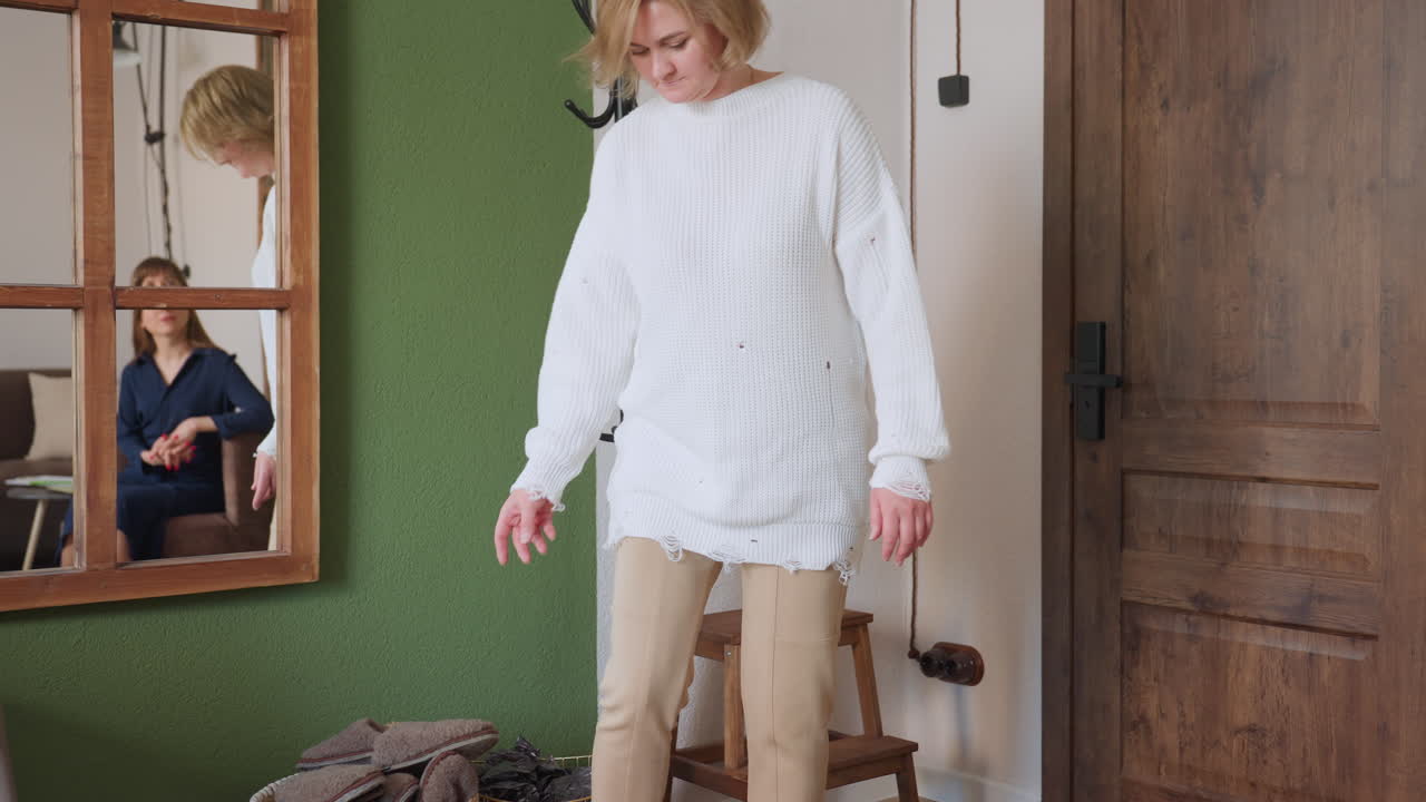 Patient wears brown crocs by side of room near door, basket and coat rack in view, while mirror on green wall reflects therapist seated on sofa calmly waiting, creating professional atmosphere