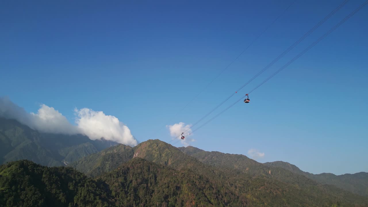 Le riprese aeree con drone catturano una funivia che scivola sulle pendici boscose delle montagne sul Fansipan nel Vietnam settentrionale con una lussureggiante copertura forestale, colline a strati e la calda luce del sole sotto un cielo azzurro limpido.