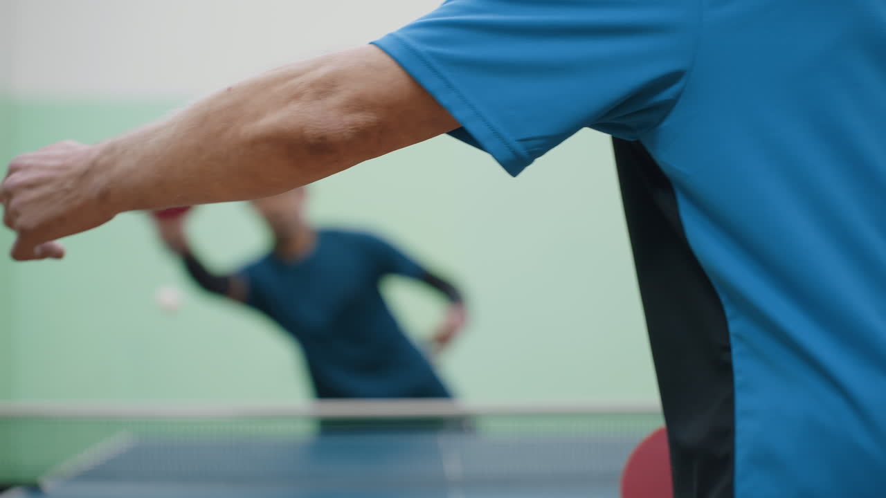 Close up back view of tennis player in blue shirt standing near table during indoor match while opponent with paddle appears blurred in background