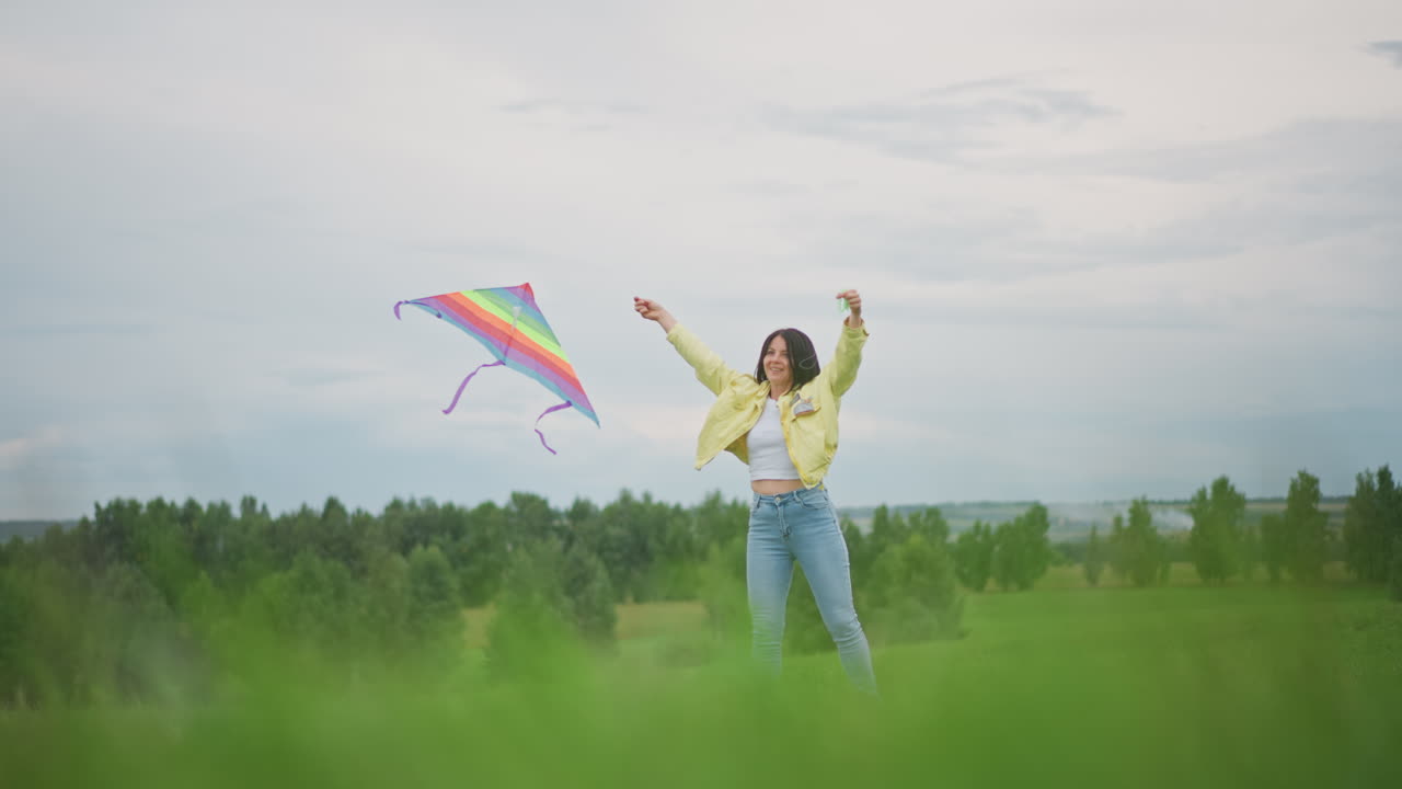 Female with vibrant kite in countryside, Lady soaring multicolored kite above green meadows in clouds, Woman enjoying bright rainbow kite flying over sprawling fields under overcast sky