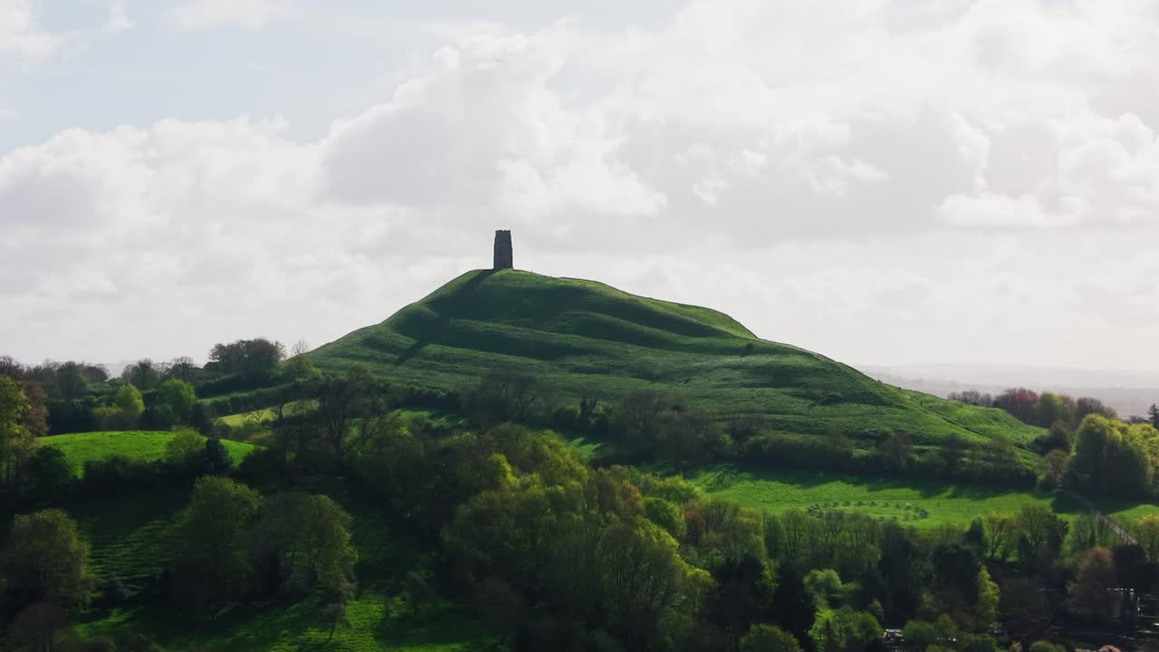 Bred luftoptagelse af Glastonbury Tor, der rejser sig over Somersets landskab, viser den ikoniske bakke og tårn omgivet af grønt landskab i Storbritannien.