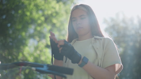 young woman stands beside her bicycle, carefully removing biker glove from her right hand with focused expression, sunlight filters through green trees, casting a soft glow on her