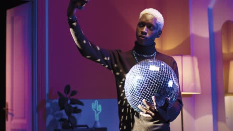 joven afroamericano con estilo sosteniendo una bola disco y haciendo selfies usando un teléfono inteligente en una fiesta retro en casa