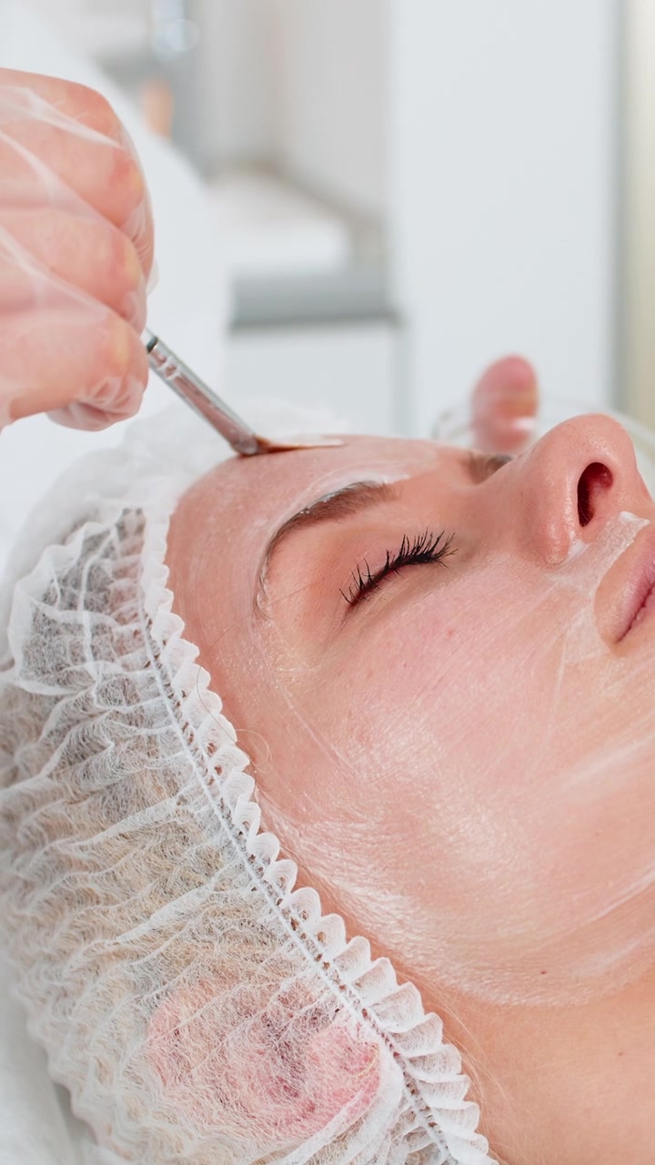 Estetista che applica una crema medica idratante sul viso di una giovane donna con dei pennelli durante una terapia enzimatica