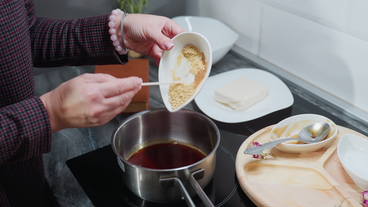 Partial view of person in checkered sleeve pouring powdered ingredients into hot oil pot on electric stove, with wooden tray, spices, and cooking utensils on marble countertop in modern kitchen