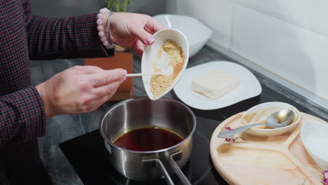 vista parcial de una persona con manga a cuadros vertiendo ingredientes en polvo en una olla de aceite caliente en una estufa eléctrica, con bandeja de madera, especias y utensilios de cocina en la encimera de mármol en la cocina moderna