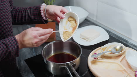 partial view of person in checkered sleeve pouring powdered ingredients into hot oil pot on electric stove, with wooden tray, spices, and cooking utensils on marble countertop in modern kitchen