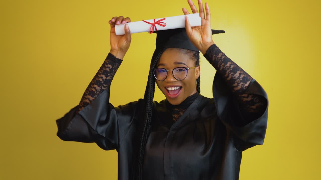 un estudiante de posgrado alegre tiene un diploma internacional y expresa felicidad y emociones positivas. estudiante con una túnica negra y sombrero sobre un fondo sólido amarillo. concepto de educación