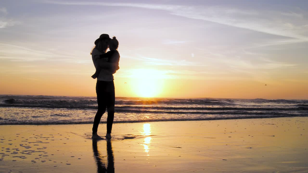 한 여성이 4k에서 해변에서 그녀의 아이와 함께 멋지고 다채로운 일몰을 즐기고 있습니다.