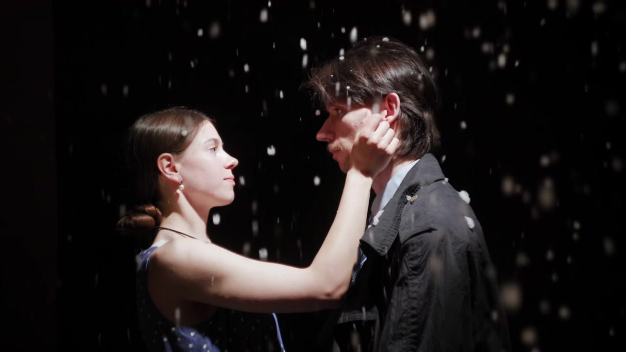 A young woman gently reaches up to touch a man's cheek in a dark studio scene with falling water, dramatic side lighting, and a black background. The two
