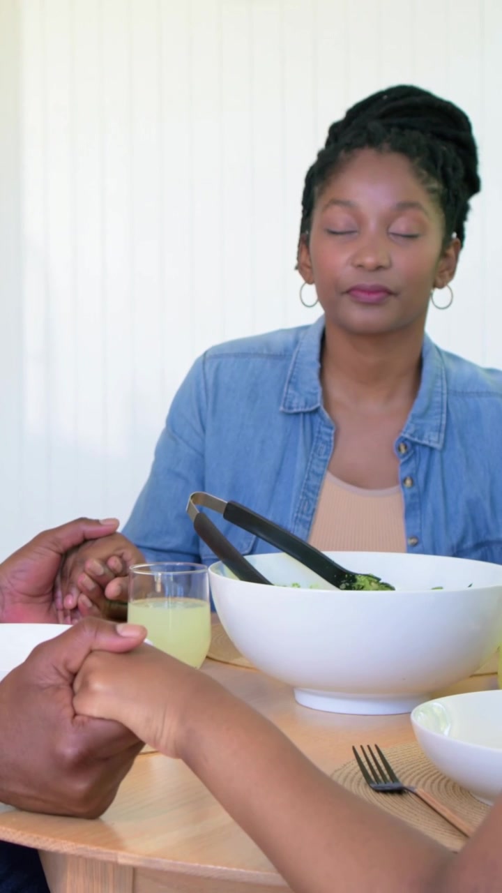 Vertical video: Holding hands woman praying at home table, man in orange serving salad with tongs