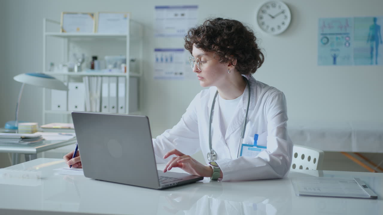 Young Female Doctor Using Laptop and Taking Notes in Medical Office