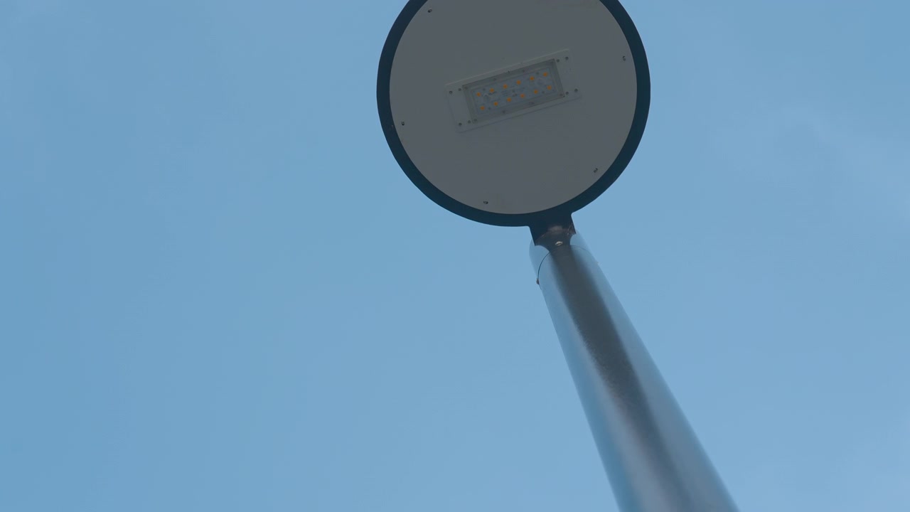 Vista de baixo ângulo de uma cabeça de poste de luz circular montada em um poste alto, centralizada contra um céu azul claro sob luz diurna suave com um fundo urbano limpo e minimalista