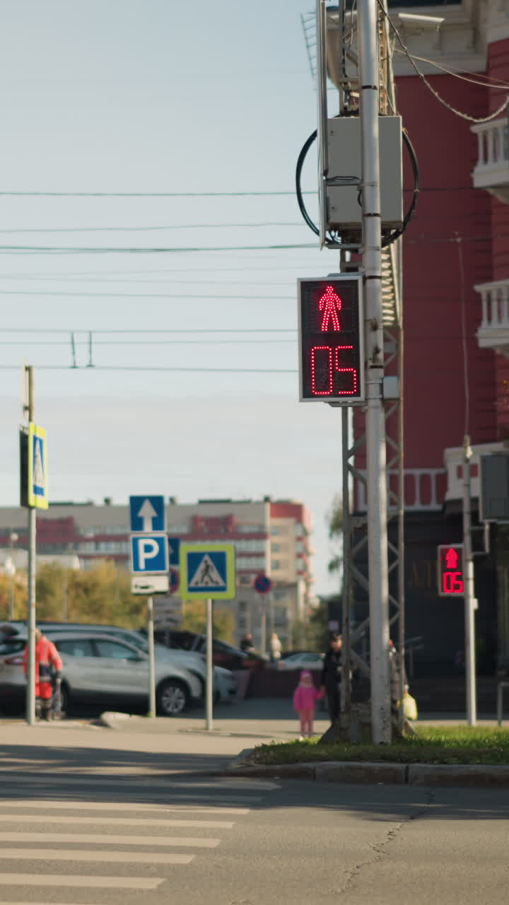 Niño esperando en el paso de peatones con el semáforo en rojo; una figura pequeña con un abrigo rosa está de pie en la acera, junto a coches aparcados y un edificio de apartamentos, mientras la señal de peatones hace la cuenta atrás; los conductores están al ralentí, a la luz del día.