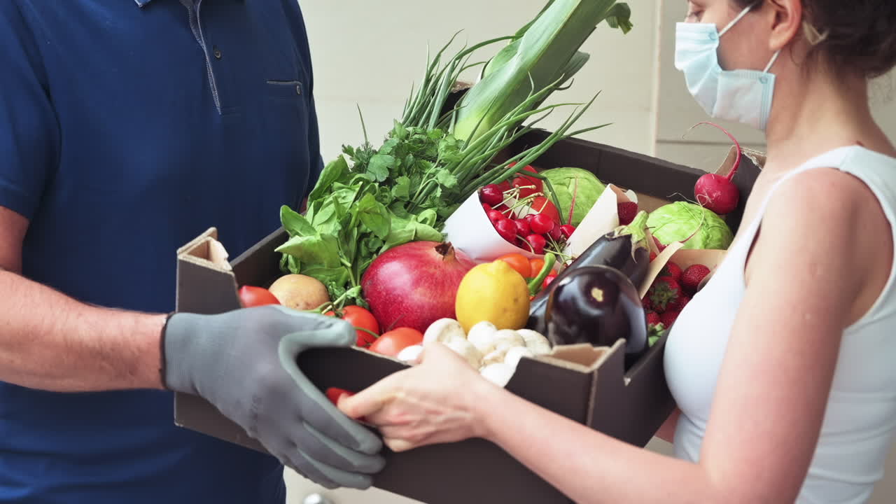 Woman in mask accepting a box of organic fruits and vegetables from delivery person