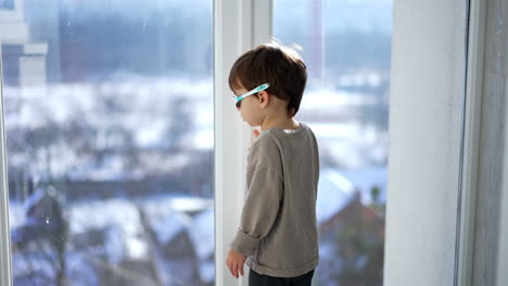 baby boy in grey sweatshirt and sunglasses stands at the panoramic window. little kid looking outdoors on sunny weather.