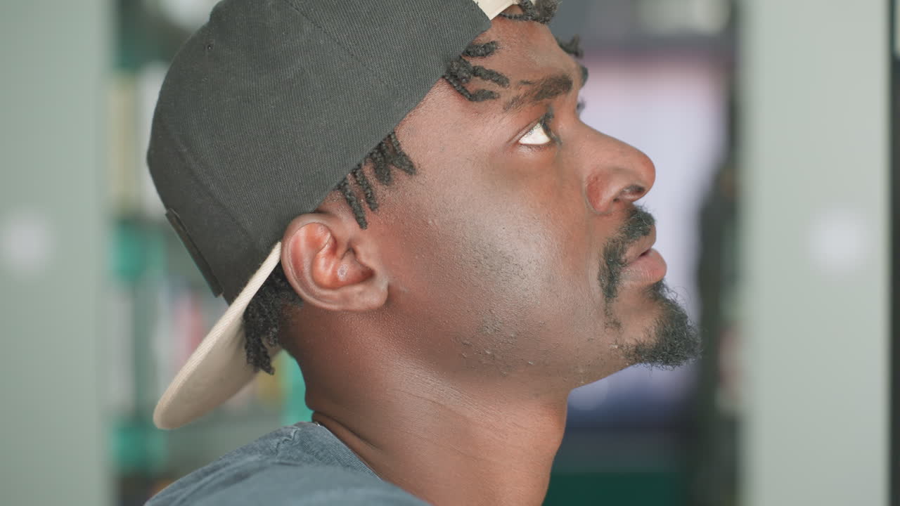 Primer plano de un joven con ropa informal, gorra al revés y gafas de sol en la camisa, mirando pensativamente mientras consulta libros en una estantería de la biblioteca en un tranquilo espacio académico