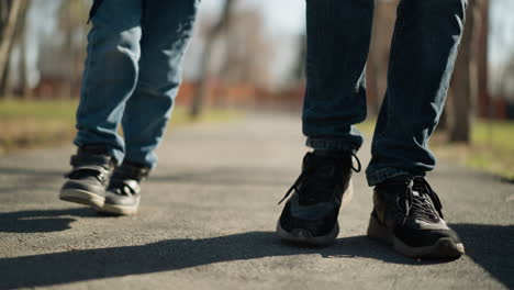 a close-up of an adult and a child wearing jeans and boots walking together on a pavement, the child is walking in an irregular pattern, with blurred view of house around