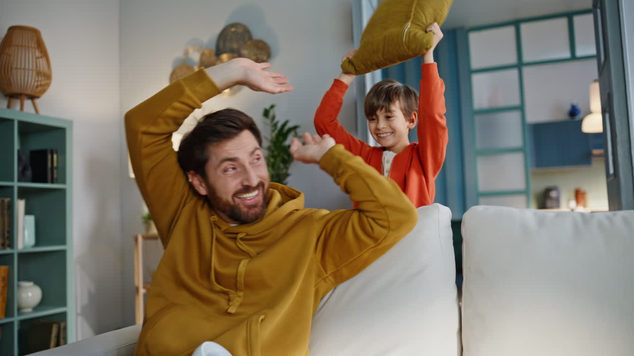 Cute son fighting pillow bearded dad sitting cozy living room couch closeup