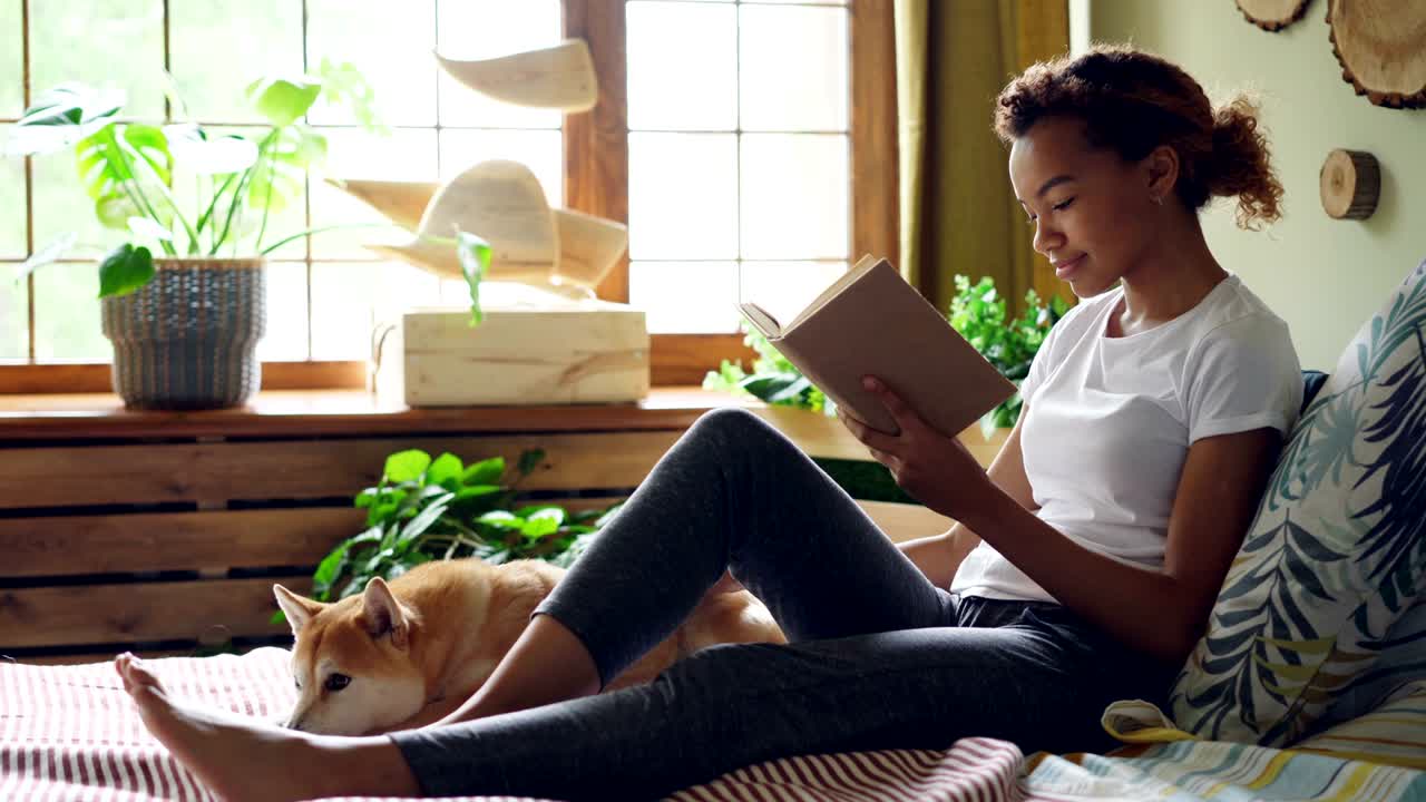 Pretty girl mixed race student is reading book and caressing pet dog expressing love and care sitting on bed in the house. Youth, hobby and animals concept.