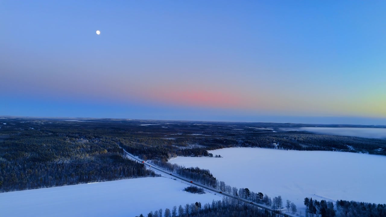 Ilmakuva moottoritiestä lumisen maiseman läpi kuun noustessa, Lapissa. Suora tie halkoo jäätyneitä peltoja ja metsää vaaleanpunaisen ja sinisen hämärätaivaan alla.