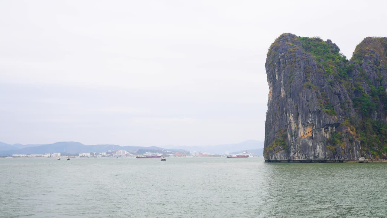 Dramatic limestone karsts with steep vertical cliffs rise from Ha Long Bay's jade-green waters, while cargo ships dot the distant horizon, creating a contrast of natural wonder and maritime activity