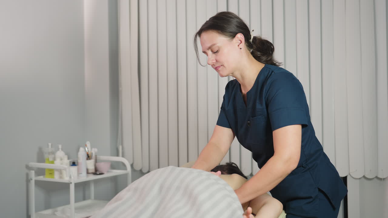 Beautician massaging back of recipient on massage bed in spa room with cream on trolley creating soothing wellness ambiance for client relaxation experience under tranquil atmosphere