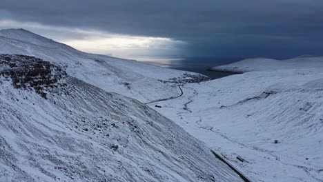 aerial drone view above mountains of small road leading into a village covered in snow with ocean